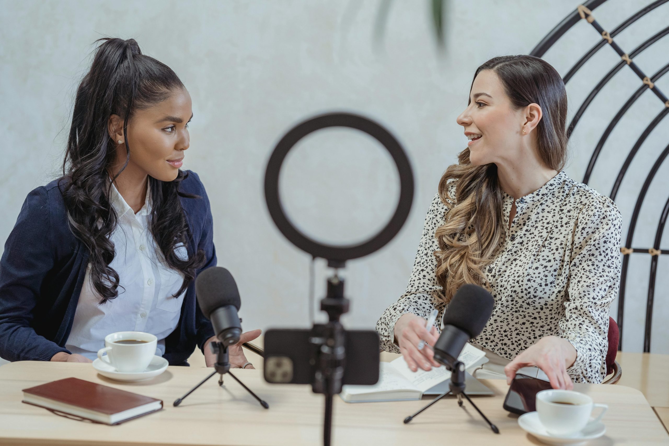 Two women sitting at a table, holding microphones, engaged in conversation over cups of coffee.